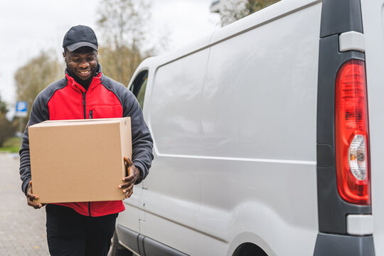 Black Young Adult Delivery Guy In Red Pullover And Black Cap Smiling Walking To White Van With Cardboard Box Parcel. Horizontal Outdoor Shot. High Quality Photo