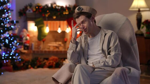 Devastated young man holding head in hands thinking sitting on the right in rocking chair. Portrait of depressed Caucasian LGBT guy at home on Christmas eve. Loneliness and holidays