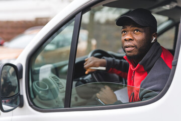 Portrait of black young adult delivery guy wearing pullover and black cap sitting inside white van leaning out the window to look for correct address. Horizontal shot. High quality photo