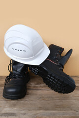 close-up of white hardhat, pair of new black work boots made of leather with reinforced cape on old wooden boards, concept of special protective professional shoes, work safety, builder's day