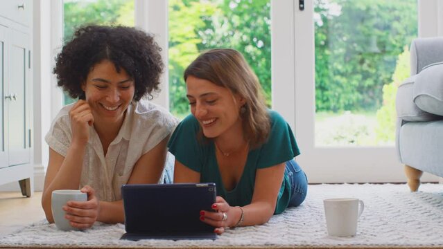 Same sex female couple or friends at home sitting in lounge using digital tablet to shop or book holiday and drinking coffee -shot in slow motion
