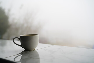 A cup of white coffee rests on the edge of a tiled balcony with a thick white mist in the background.
