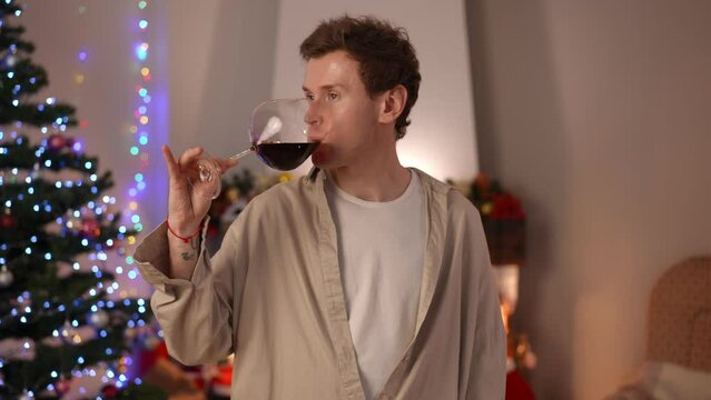 Confident man looking away talking drinking red wine from glass standing in living room on Christmas eve. Portrait of young Caucasian LGBT guy celebrating New Year alone at home. Solitude concept