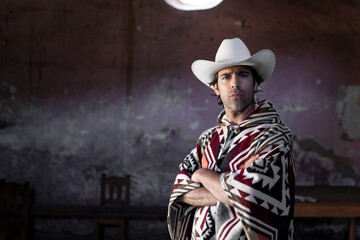 A young Mexican is posing with crossed arms wearing a hat and a poncho