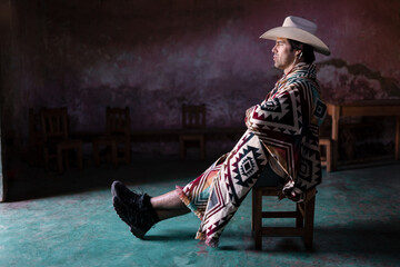 A young Mexican is sitting with crossed arms inside of a church wearing a hat and a poncho - obrazy, fototapety, plakaty