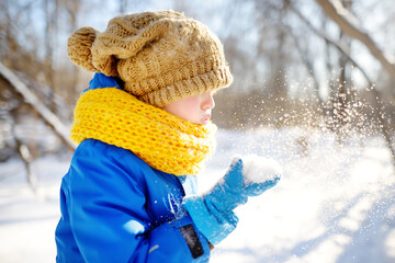 Little boy blowing snow from his hands. Child enjoy walking in the park on snowy day. Baby having...