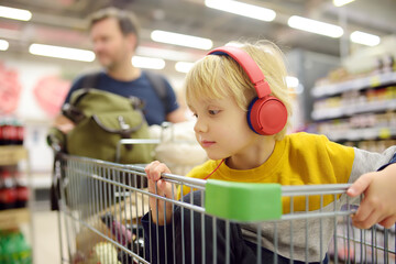 Cute preschooler boy with headphone and player is sitting in a shopping cart at a food store or...