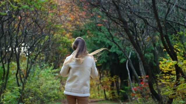Dark-haired lady in white jacket walking away by the path through the forest. Romantic woman distancing from camera in autumn park.