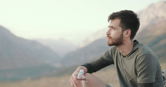 Portrait of an ethnic Caucasian with an inspired bearded face. a young adult thoughtful guy is sitting on top of a mountain, drinking water from a plastic bottle and dreaming, enjoying the sunset rays