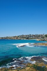 Sydney eastern suburbs headland, cloudless blue skies and waves crashing against the rocks, as seen from the Bondi to Tamarama coastal walk — Tamarama, Sydney; New South Wales, Australia