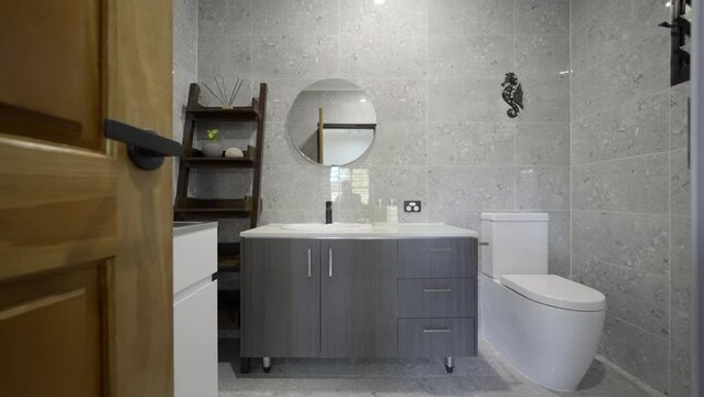 Luxurious modern bathroom with sleek grey tiles white bath and white fixtures in a contemporary residential home. Round mirror. Wooden door wooden shelving. White bench top cupboard. glass shower door
