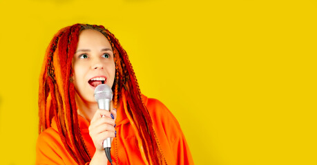 Young woman with bright dreadlocks singing into microphone on yellow background. Emotional female...