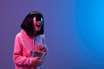 Laughing excited awesome brunet woman in pink hoodie trendy specular sunglasses eat popcorn look aside posing isolated in blue violet color light background. Neon party Cinema concept. Copy space