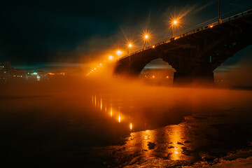 a bridge over an icy river at winter night. the snow-covered bank of the winter river at night, from which steam rises, and the light of lanterns. Bridge over the river. Selective focus.
