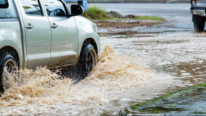 Pickup truck passing through flooded road. Driving car on flooded road during flood caused by torrential rains. Flooded city road with large puddle. Splash by car through flood water. Selective focus. - obrazy, fototapety, plakaty