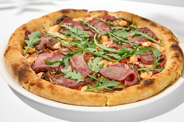 Italian pizza with roast beef, arugula and spicy sauce on light background with shadows. Meat pizza with sliced roast beef on white table. Italian food in trendy menu restaurant