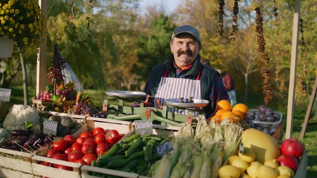 Senior farmer stands at the stall with fresh colorful fruits and vegetables, looks at camera. Weekend shopping at local farmers market outside. Vegetarian, organic and healthy food. Agriculture.