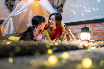 Asian mother and cute smiling girl reading a book together while lying in illuminated tent in kid...