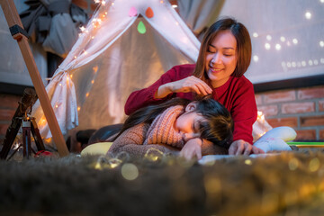 Asian mother taking care of her cute girl to sleep while lying in illuminated tent in kid bedroom. Cheerful ethnic woman and lovely daughter playing under a cozy hut.
