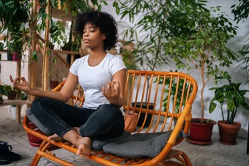 Tuinposter Serene calm black young woman sitting with closed eyes on sofa in lotus pose, practicing yoga meditation at home, relaxing and breathing deep for mental health in room with biophilia design. © dikushin