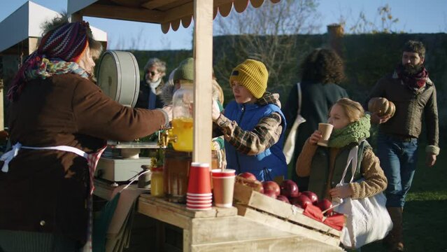 Elderly woman at the stall pours lemonade or tea into paper cups for boy and girl. People relaxing on fresh air on background. Spending weekend at local farmers market or autumn fair outdoors.