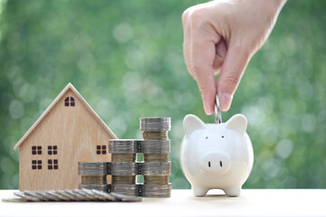 Woman hand putting a coin into piggy bank with model house on nature green background,Save money...