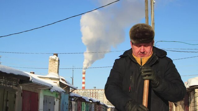 A man in a warm jacket is resting leaning on a wooden snow shovel. Large snowdrifts around after a snowstorm, sunny day. City life, heavy snowfall. UHD 4K.
