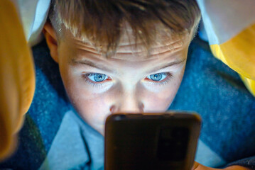 Serious boy under the blanket at night in his bed watching prohibited content.Child gadget...