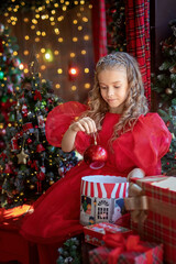 Cute little girl in a red dress in a New Year's interior with a Christmas tree holding a red ball...