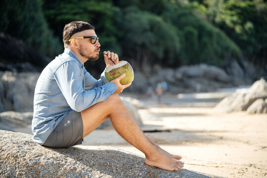 Healthy food in the morning..A man drinks fresh coconut milk on the beach in the morning barefoot while sitting on a stone.