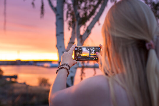 Frau fotografiert Sonnenuntergang am Ufer mit Birke im Hintergrund. Blonde Frau fotografieren mit Mobiltelefon.