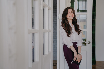 Cheerful brunette woman in white shirt carrying diary toothy smiling looks aside happily, completed her work. Attractive Hispanic teacher comes in classroom, smiling office manager starts day.