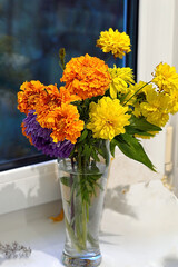 Bouquet of orange marigolds, blue aster and yellow flowers in a glass vase. The bouquet stands on a white window sill by the window. Top side view.