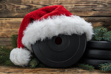 Heavy dumbbells barbell weight plate disc in a red Santa Claus Christmas hat. Healthy fitness lifestyle holiday season winter composition. Gym workout and sport training concept, with tree branches.