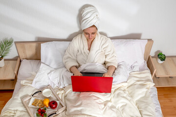 Woman working with laptop, eating fruit breakfast in bed at home or hotel in the morning. Lady...