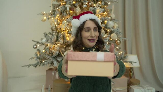 Woman wearing green sweater and Santa's cap presenting a gift near the tree