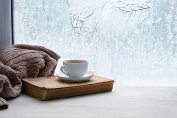 tea cup, book, sweater and winter frozen window. cozy mood, home comfort in snowy cold weather....