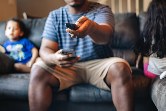 Father At Home Sitting On Couch With Kids In The Background While Holding Tv Remote And Cellphone