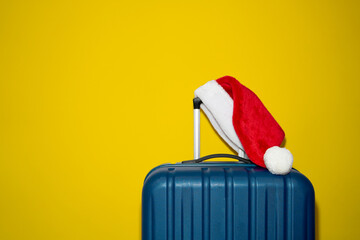 Blue plastic suitcase with Santa Claus cap over yellow background. Concept of travel and visiting relatives on Christmas Holidays