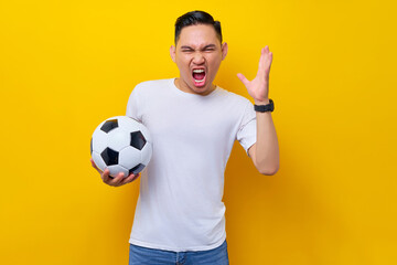 Screaming upset angry young Asian man football fan in a white t-shirt holding a soccer ball and yelling raised hand isolated on yellow background. People sport leisure lifestyle concept
