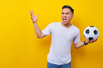 Screaming upset angry young Asian man football fan in a white t-shirt holding a soccer ball and...