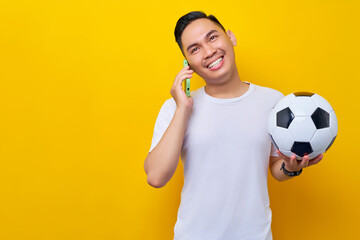 Smiling young Asian man football fan wearing a white t-shirt holding a soccer ball and talking on a...