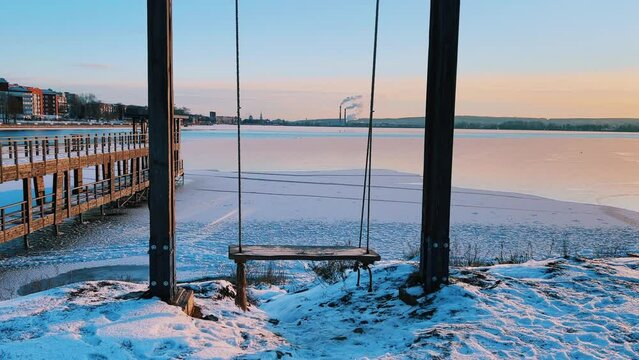 An empty wooden swing moving outdoors, without people. Beautiful view of the pond covered with snow and ice against the background of the smoking pipes of the Topol-M rocket factory. Russia