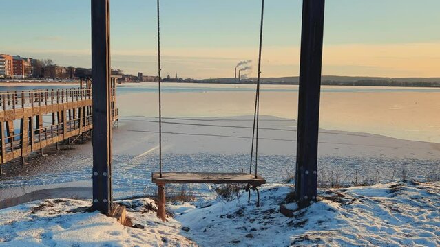 An empty wooden swing moving outdoors, without people. Beautiful view of the pond covered with snow and ice against the background of the smoking pipes of the Topol-M rocket factory. Russia