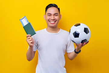 excited young Asian man football fan wearing a white t-shirt holding a soccer ball and passport ticket boarding pass isolated on yellow background. People sport leisure lifestyle concept