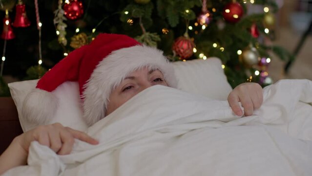 adult female in red Santa hat waking up in Christmas morning in bed near decorated Christmas tree opening warm white blanket from face, smiling , stretching up festive holiday morning