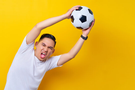 Cheerful Young Asian Man Footbal Fan In White T-shirt Cheer Up Support Favorite Team Catching Soccer Ball Gesture Isolated On Yellow Background. People Sport Leisure Lifestyle Concept