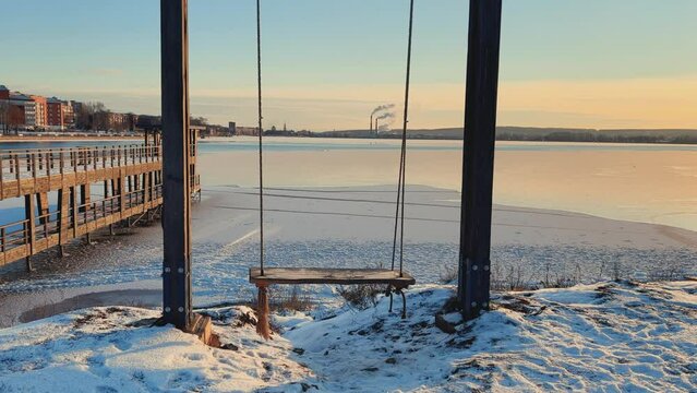 An empty wooden swing moving outdoors, without people. Beautiful view of the pond covered with snow and ice against the background of the smoking pipes of the Topol-M rocket factory. Russia