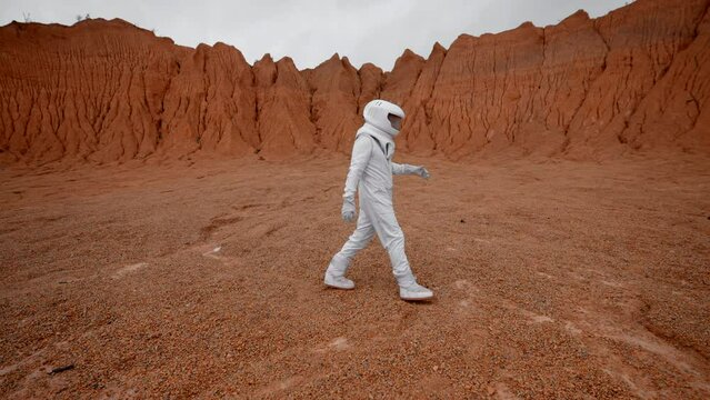 Sideview of an astronaut walking by orange surface of unknown far planet. Somewhere in space, humans colonise planet and start to explore surface. Wide shot in slow motion.