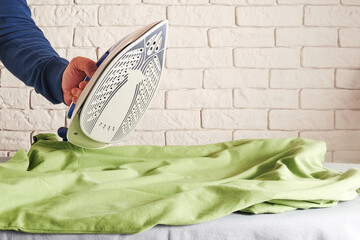 Man hand holding hot iron and smooths wrinkled green shirt. Household work and laundry theme. Copy space for text
